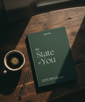 En mørkegrønn journal med tittelen "The State of You" ligger på et trebord ved siden av en kopp kaffe og en penn, som fremhever klarhet og balanse.