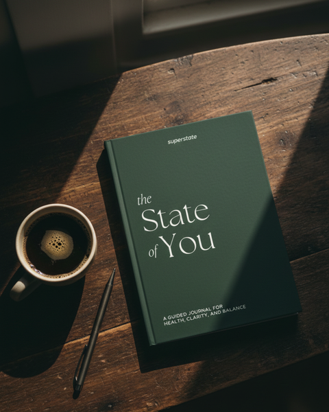 En mørkegrønn journal med tittelen "The State of You" ligger på et trebord ved siden av en kopp kaffe og en penn, som fremhever klarhet og balanse.